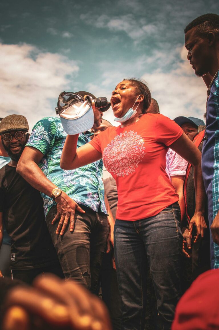 A passionate woman addresses a lively crowd outdoors using a microphone on a sunny day.